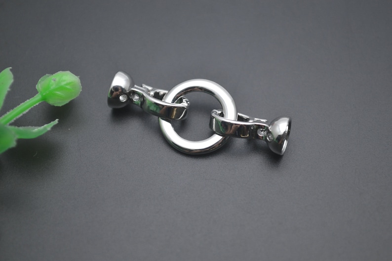 May include: A polished, silver-coloured clasp featuring a central circular ring. The clasp has two rounded ends with a hinged mechanism for opening and closing. Likely used for jewellery or accessories.