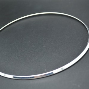 May include: A silver-colored choker necklace with a simple, minimalist design. The necklace is a thin, smooth band that forms a complete circle, with a clasp closure. The jewelry is displayed against a dark background.