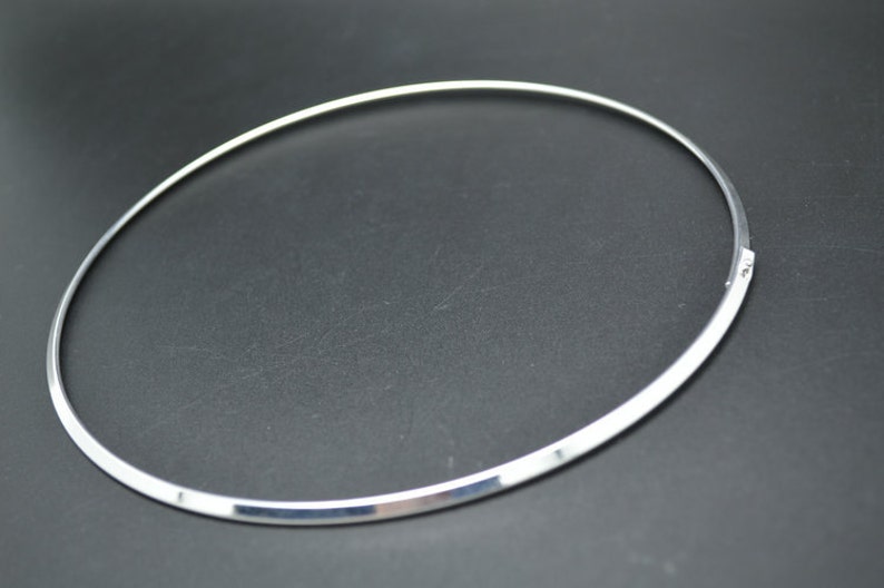 May include: A silver-colored choker necklace with a simple, minimalist design. The necklace is a thin, smooth band that forms a complete circle, with a clasp closure. The jewelry is displayed against a dark background.