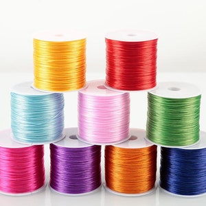 Könnte beinhalten: Sortiment von acht Spulen mit farbigem elastischem Garn, jede um eine weiße Kunststoffspule gewickelt. Die Farben sind gelb, rot, hellblau, rosa, grün, lila, orange und dunkelblau.