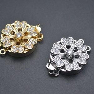 May include: Two flower-shaped clasps for jewelry making. One is gold with clear rhinestones, the other is silver with clear rhinestones.