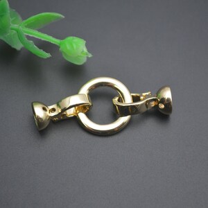 May include: A gold-coloured jewellery clasp with a circular ring and two end pieces. The clasp is made of metal and has a shiny finish. The clasp is designed to connect two pieces of jewellery together. A green plant is in the background.