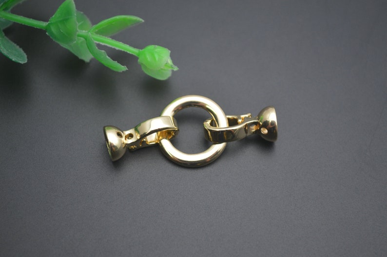 May include: A gold-coloured jewellery clasp with a circular ring and two end pieces. The clasp is made of metal and has a shiny finish. The clasp is designed to connect two pieces of jewellery together. A green plant is in the background.