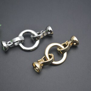 May include: Two metal clasp connectors, one silver and one gold, are displayed against a dark background. Each clasp features a circular ring connected to a decorative, rounded end piece. The clasps are designed for jewellery making or similar crafts.