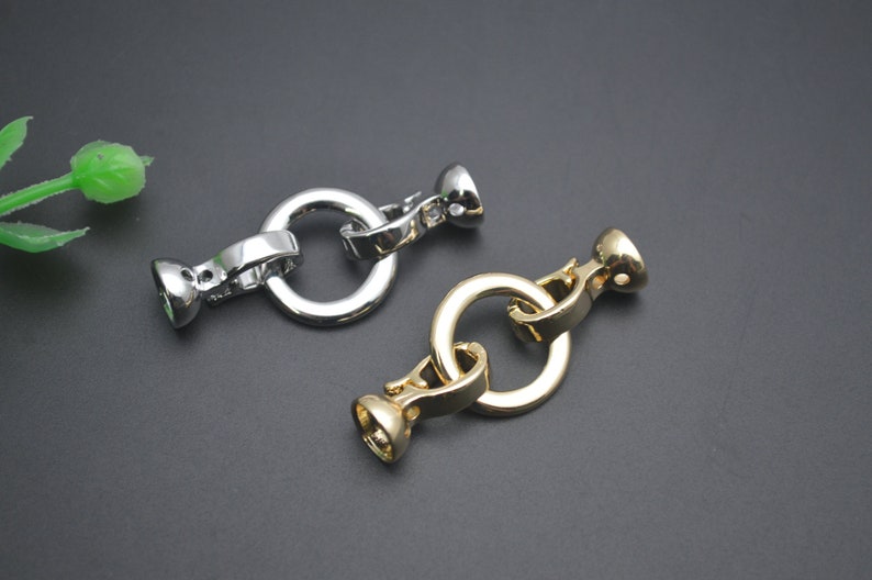 May include: Two metal clasp connectors, one silver and one gold, are displayed against a dark background. Each clasp features a circular ring connected to a decorative, rounded end piece. The clasps are designed for jewellery making or similar crafts.