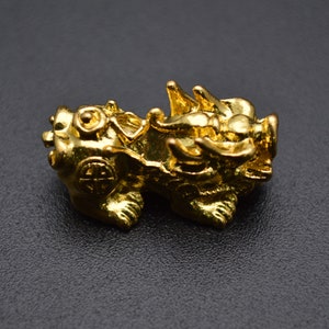 May include: A gold-colored metal charm in the shape of a mythical Chinese dragon. The dragon is depicted in a crouching position with its head turned to the side. The charm has intricate details and a shiny finish.