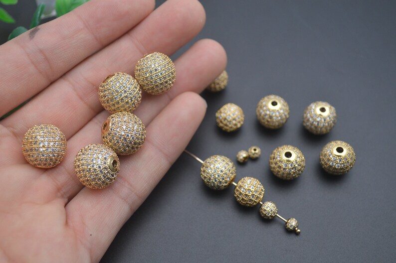 May include: A collection of gold-toned round beads with clear crystal accents. The beads have a hole in the center for stringing.
