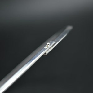 May include: Close-up of a silver metal tool with a pointed tip and a small, round rivet. The tool has a flat, rectangular section with a small, raised area. The background is a dark gray.