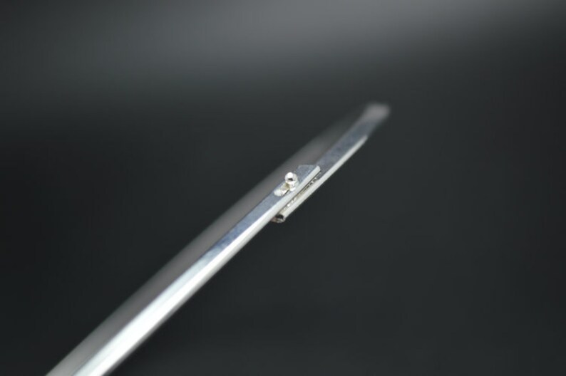 May include: Close-up of a silver metal tool with a pointed tip and a small, round rivet. The tool has a flat, rectangular section with a small, raised area. The background is a dark gray.
