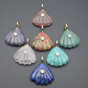 May include: A collection of seven gemstone pendants in various colors, including purple, pink, green, and blue. Each pendant has a shell-like design with a gold bail and a small, round, clear stone in the center. The pendants are displayed on a gray surface.