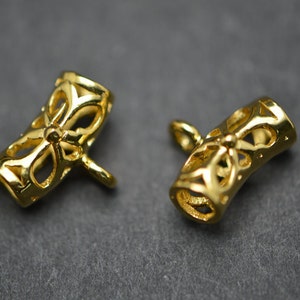 May include: Two gold-colored, open-work jewelry clasps. Each clasp is cylindrical with a floral design and a small loop extending from one end. The clasps are on a dark gray surface.