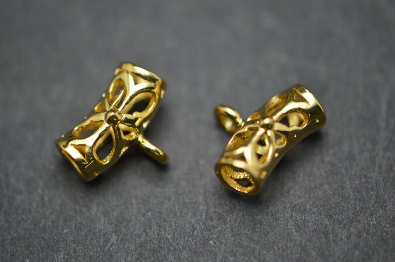 May include: Two gold-colored, open-work jewelry clasps. Each clasp is cylindrical with a floral design and a small loop extending from one end. The clasps are on a dark gray surface.