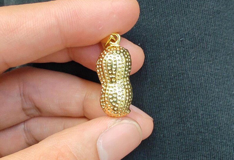 May include: A gold-colored peanut charm pendant. The peanut is textured with small bumps and has a shiny finish.