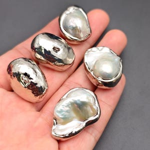 May include: Five irregular shaped, white, baroque pearls with a silver sheen. The pearls are on a hand.