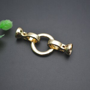 May include: A gold-coloured jewellery clasp with a circular ring and two end pieces. The clasp is made of metal and has a shiny finish. The clasp is designed to connect two pieces of jewellery together.