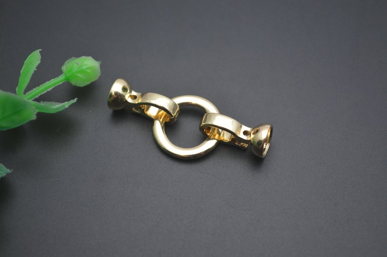May include: A gold-coloured jewellery clasp with a circular ring and two end pieces. The clasp is made of metal and has a shiny finish. The clasp is designed to connect two pieces of jewellery together.