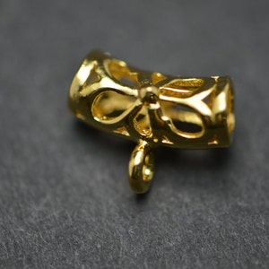 May include: A gold-colored jewelry component with a bow-shaped design. The component has a cylindrical shape with openwork patterns and a small loop at the bottom for attachment. The metal has a shiny finish.