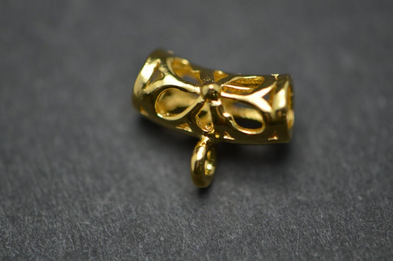 May include: A gold-colored jewelry component with a bow-shaped design. The component has a cylindrical shape with openwork patterns and a small loop at the bottom for attachment. The metal has a shiny finish.