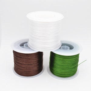 May include: Three spools of elastic cord in white, brown, and green. The spools are made of white plastic and have a clear plastic center.