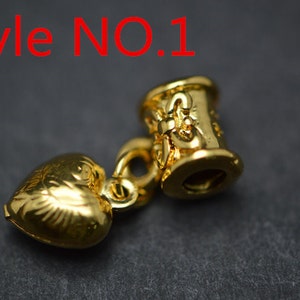 Puede incluir: Encanto dorado con un colgante en forma de coraz&oacute;n y una cuenta cil&iacute;ndrica con dise&ntilde;os intrincados. La cuenta tiene el texto "Style NO. 1" en ella.