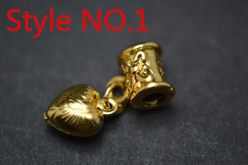 Puede incluir: Encanto dorado con un colgante en forma de coraz&oacute;n y una cuenta cil&iacute;ndrica con dise&ntilde;os intrincados. La cuenta tiene el texto "Style NO. 1" en ella.