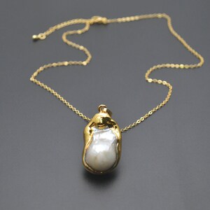 May include: A gold chain necklace with a large, irregular pearl pendant. The pearl is set in a gold frame and has a white, iridescent sheen.