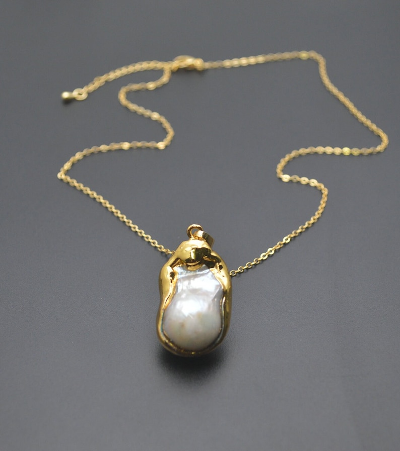 May include: A gold chain necklace with a large, irregular pearl pendant. The pearl is set in a gold frame and has a white, iridescent sheen.