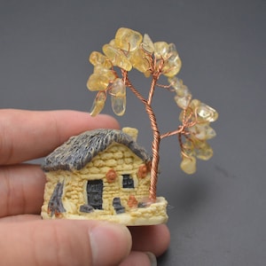 Könnte beinhalten: Ein kleines Steinhaus mit Strohdach und einem drahtumwickelten Baum mit gelben Edelsteinen als Blätter. Das Haus hat eine schwarze Tür und Fenster.