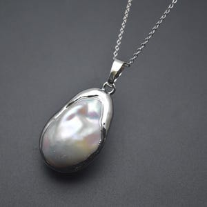 May include: A silver chain necklace with a large, white baroque pearl pendant. The pearl is set in a silver bezel and has an irregular shape.