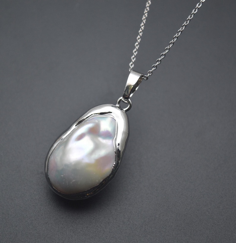 May include: A silver chain necklace with a large, white baroque pearl pendant. The pearl is set in a silver bezel and has an irregular shape.