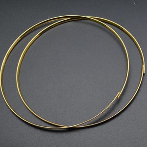 May include: Two gold-colored, thin, circular necklaces are displayed against a dark background. One necklace is slightly overlapping the other. The necklaces appear to be made of metal and have a clasp closure.