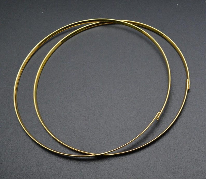 May include: Two gold-colored, thin, circular necklaces are displayed against a dark background. One necklace is slightly overlapping the other. The necklaces appear to be made of metal and have a clasp closure.