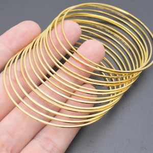 May include: A collection of gold-colored bangles, each a thin, smooth circle. The bangles are stacked together, creating a layered effect. The jewelry is held in a hand against a dark gray background. The bangles are a bright, shiny gold.