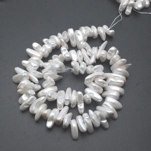May include: A strand of white, irregular freshwater pearls. The pearls are strung on a white string, forming a necklace. The pearls have a lustrous, pearlescent sheen, and vary in size and shape. The background is grey.