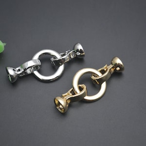May include: Two metal jewellery clasps. One is silver, the other is gold. Each clasp has a circular ring connected to two decorative end pieces. The clasps are designed to connect jewellery pieces.