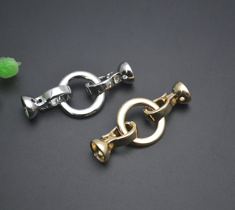 May include: Two metal jewellery clasps. One is silver, the other is gold. Each clasp has a circular ring connected to two decorative end pieces. The clasps are designed to connect jewellery pieces.