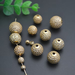 May include: A collection of gold-toned round beads with clear rhinestones. The beads are of various sizes and are arranged on a dark surface.