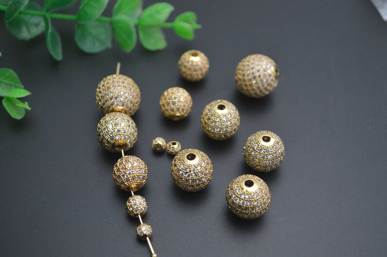 May include: A collection of gold-toned round beads with clear rhinestones. The beads are of various sizes and are arranged on a dark surface.