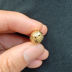 May include: A gold-colored bead with a textured surface and two small holes. The bead is round and has a slightly irregular shape.