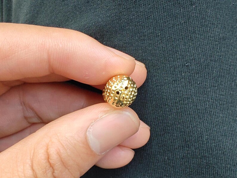May include: A gold-colored bead with a textured surface and two small holes. The bead is round and has a slightly irregular shape.