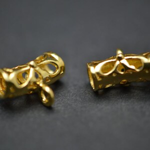 May include: Two gold-colored, cylindrical jewelry components with intricate cut-out designs. Each component has a loop at one end and a decorative pattern around the middle. The components are displayed against a dark background.