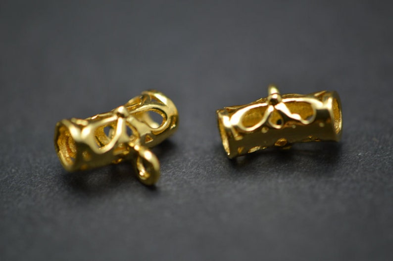 May include: Two gold-colored, cylindrical jewelry components with intricate cut-out designs. Each component has a loop at one end and a decorative pattern around the middle. The components are displayed against a dark background.