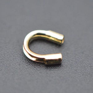 May include: A gold-coloured, U-shaped metal clasp. The clasp has a rounded curve and two straight, cylindrical ends. The surface of the clasp is smooth and reflective, with a slight sheen. The background is a dark, textured surface.