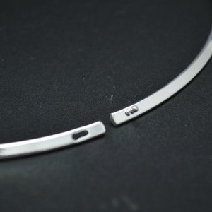 May include: A silver-colored choker necklace with a simple, minimalist design. The necklace is open, showing the clasp mechanism. The metal has a polished, reflective surface. The background is a dark, solid color.