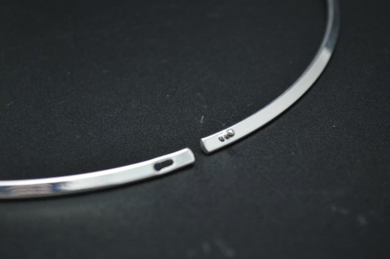 May include: A silver-colored choker necklace with a simple, minimalist design. The necklace is open, showing the clasp mechanism. The metal has a polished, reflective surface. The background is a dark, solid color.