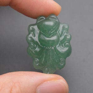 May include: A translucent green gemstone frog pendant. The frog is carved with detailed features and a decorative border. The pendant is held between fingers against a gray background. The frog pendant is a unique piece of jewelry.