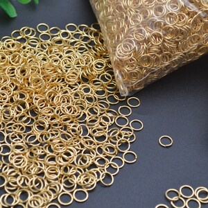 以下が含まれることがあります： 2 つの透明なプラスチック袋に入った、大量の金色のジャンプリング。ジャンプリングは小さく丸く、ジュエリー作りに最適です。