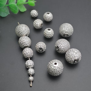 May include: A collection of silver round beads with clear rhinestones. The beads are of various sizes and are arranged on a gray surface.