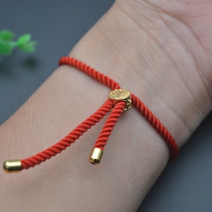 May include: A red string bracelet with a gold adjustable clasp. The bracelet is tied in a knot and has a gold charm with a design on it.