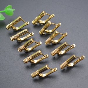 Op de afbeelding: Een verzameling goudkleurige oorringen. Elke oorring heeft een rechthoekige staaf met een getextureerd, met edelstenen bezet uiteinde en een scharniersluiting. De oorbellen zijn geplaatst op een donkergrijze ondergrond.